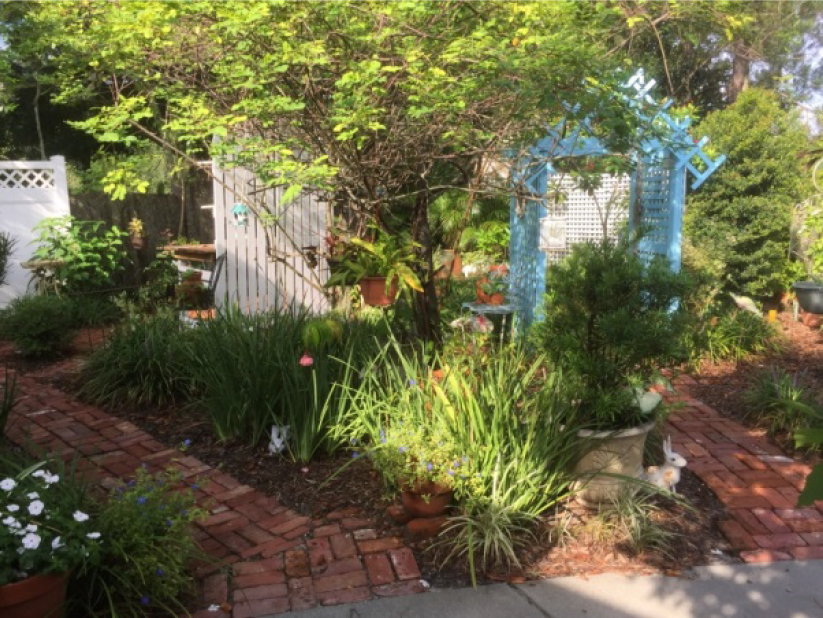 A lush garden featuring a brick pathway, green plants, flowers, and a blue trellis under a tree.