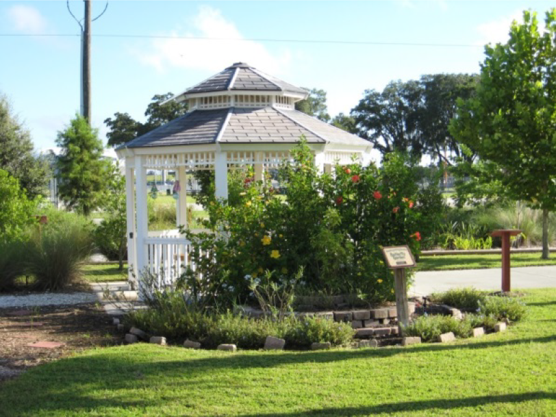 A white gazebo surrounded by colorful flowers and shrubs, set in a green park with trees in the background.