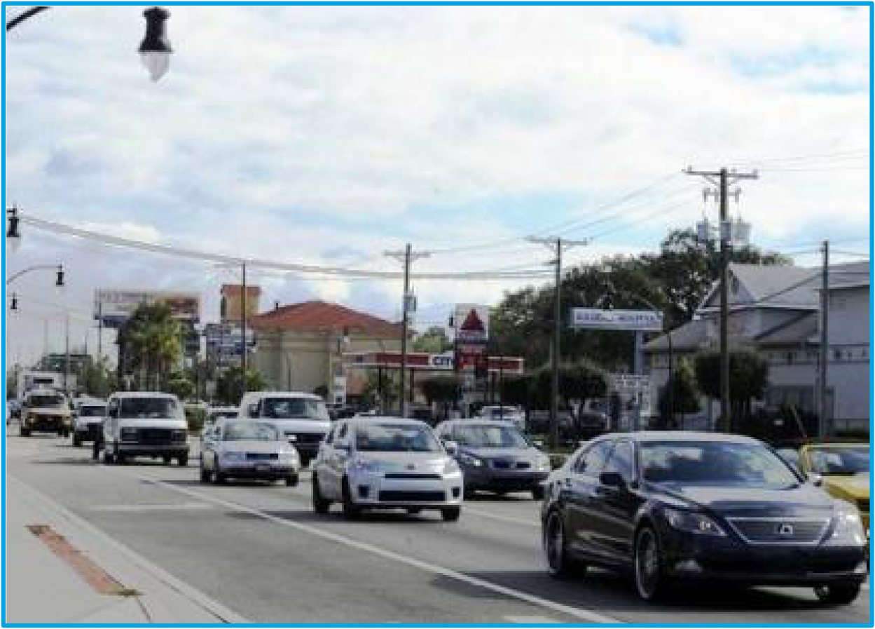 Busy street scene with several cars driving, traffic lights, and buildings lining the side of the road under a cloudy sky.