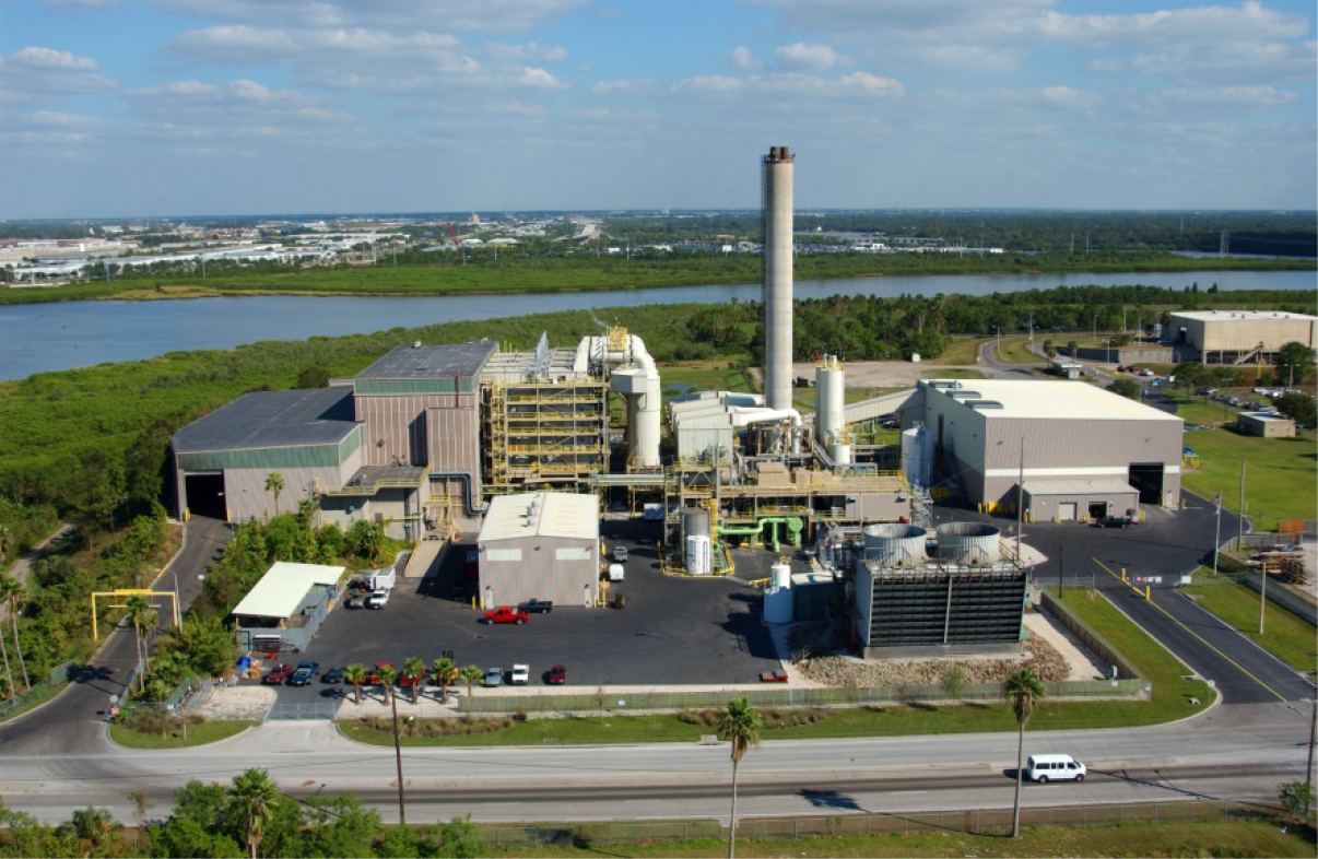 Aerial view of an industrial plant with large structures, a tall smokestack, parking area, and green surroundings near a body of water.