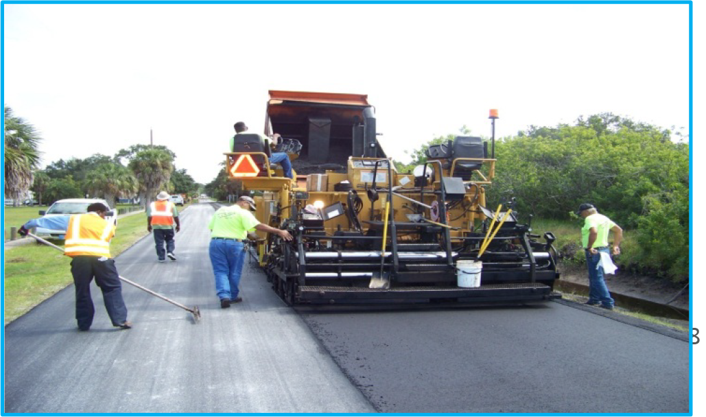 Construction workers operating a paving machine while laying asphalt on a road, with palm trees in the background.
