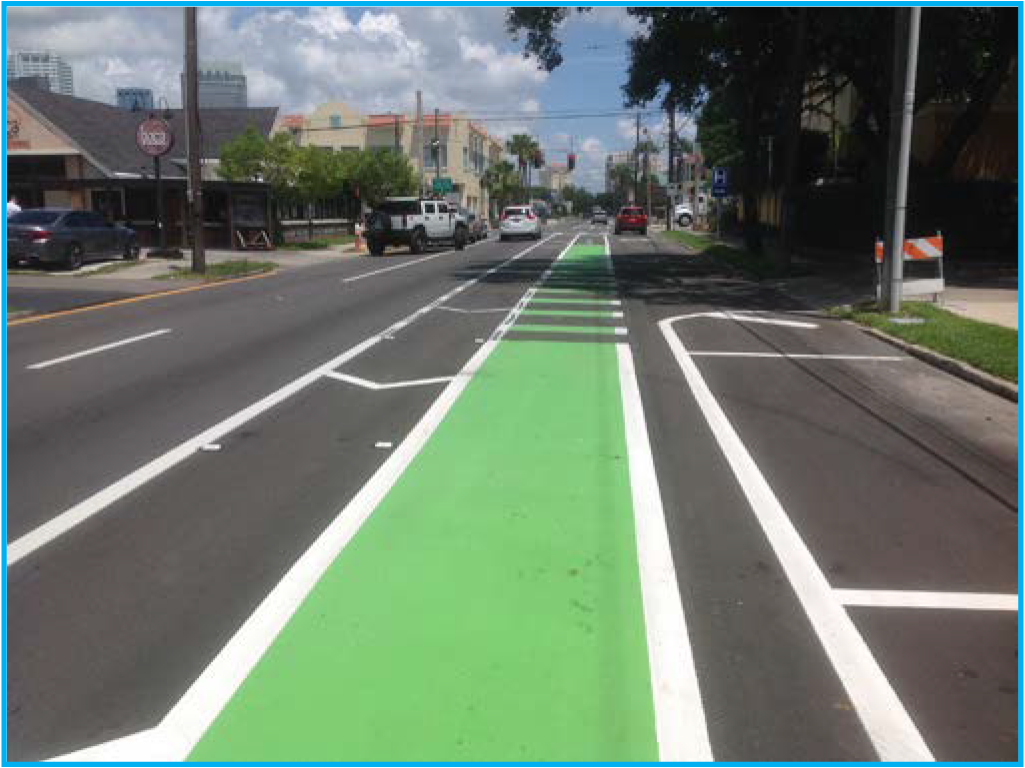 A street featuring a bright green bike lane alongside vehicle lanes, with buildings and trees visible in the background.