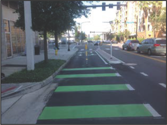 A city street featuring green bicycle lanes and a pedestrian crosswalk, with parked cars and buildings in the background.