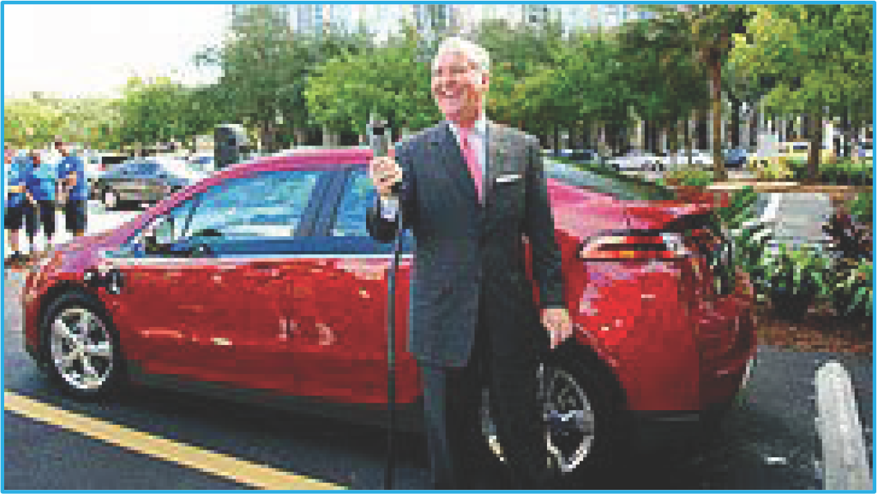 A man in a suit poses with a cane next to a red car in a parking lot surrounded by greenery.