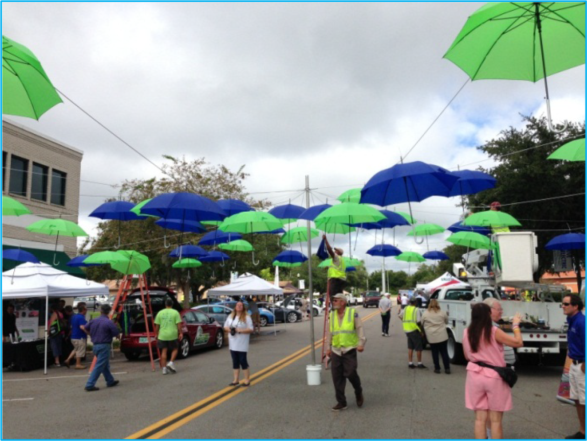 Colorful umbrellas hanging above a street with vendors and people walking in a festive outdoor market.
