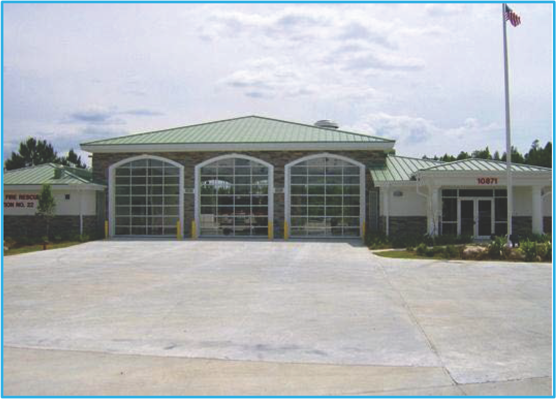 A modern fire station with a green metal roof, large glass garage doors, and an American flag outside.