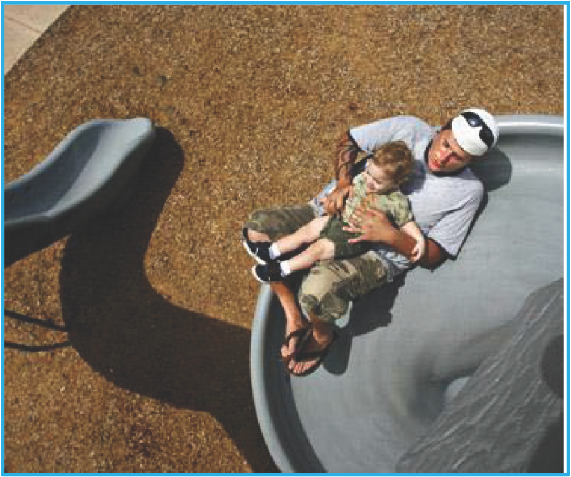 A man and a child sit together on a playground slide, with a textured ground beneath them.