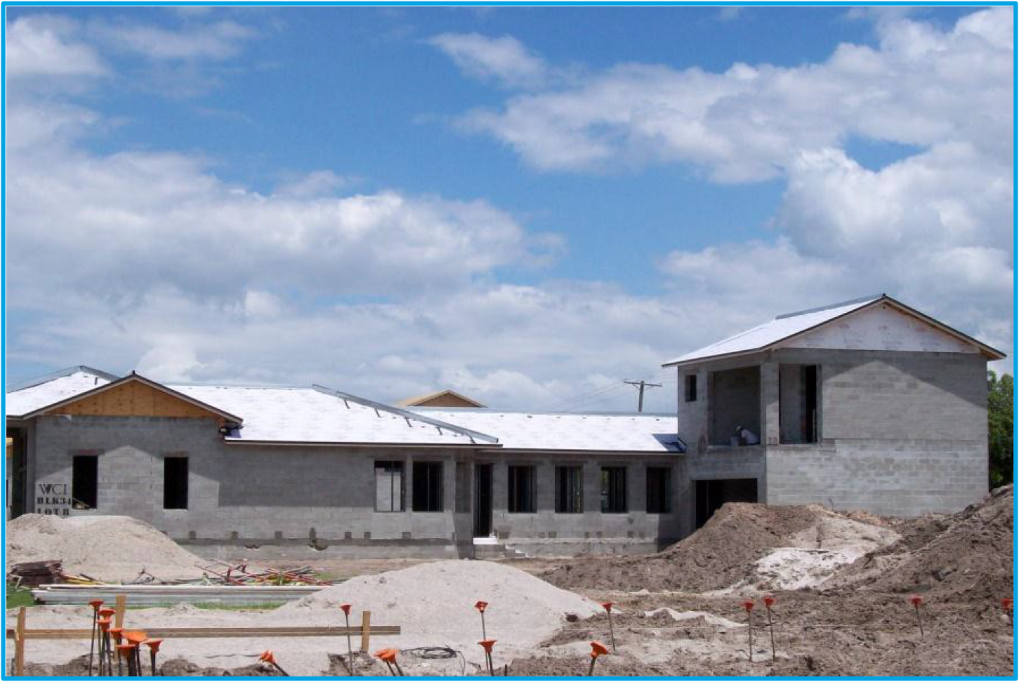 Construction site with two buildings in progress, featuring unfinished walls, a sandy area, and a cloudy sky in the background.